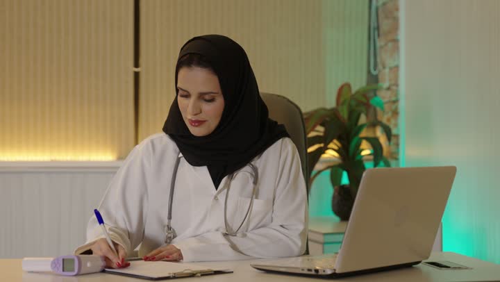 إدارة شؤون الأعمال عبر جهاز تقني حديث ومتطور، العمل فى القطاع الصحي،  طبيبة عربية خليجية سعودية ترتدي المعطف الابيض والسماعه تقوم باستخدام الحاسوب المحمول   ، مفهوم الطب والرعاية الصحية، العيادة الطبية في السعودية