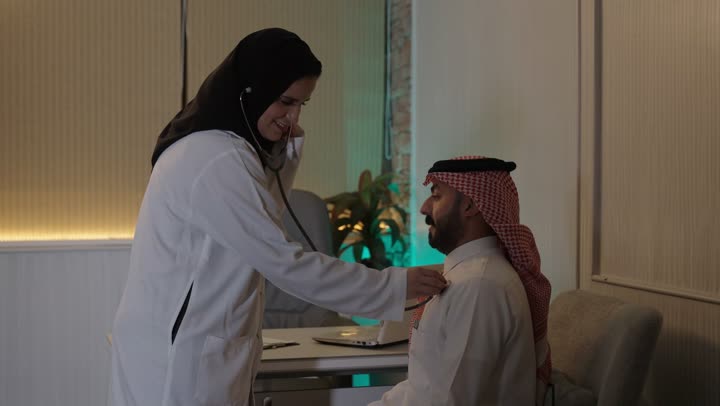 استخدام سماعة الطبيب لسماع نبضات القلب، العمل في القطاع الصحي في السعودية، القيام بالرعاية الصحية، الاهتمام بصحة المرضى، طبيبة عربية خليجية سعودية ترتدي عباءة ومئزر أبيض تقوم بفحص رجل سعودي عبر سماعة الأذن، الاهتمام بالصحة الجسدية