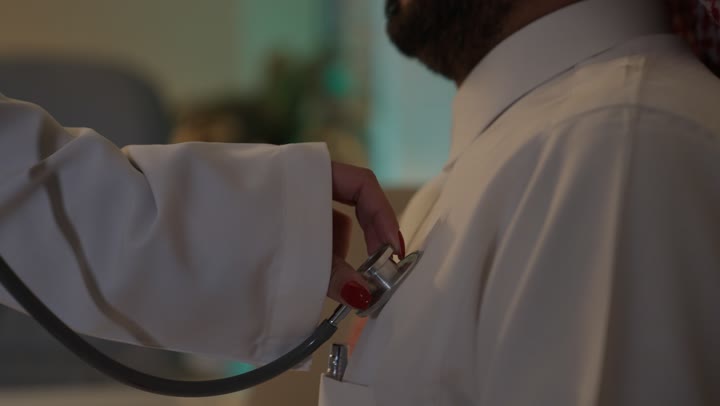 Using a stethoscope to listen to heartbeats, working in the healthcare sector in Saudi Arabia, providing healthcare, caring for patients' health, an Arab Gulf Saudi female doctor wearing an abaya and a white apron examining a Saudi man through an ear stethoscope, focusing on physical health.
