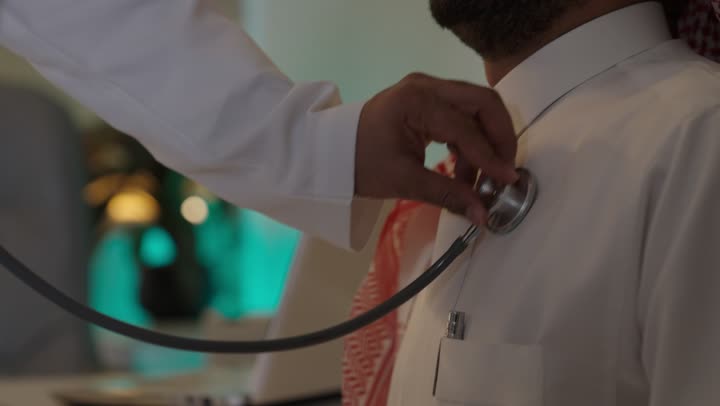 استخدام سماعة الطبيب لسماع نبضات القلب، العمل في القطاع الصحي في السعودية، القيام بالرعاية الصحية، الاهتمام بصحة المرضى، طبيبة عربية خليجية سعودية ترتدي عباءة ومئزر أبيض تقوم بفحص رجل سعودي عبر سماعة الأذن، الاهتمام بالصحة الجسدية