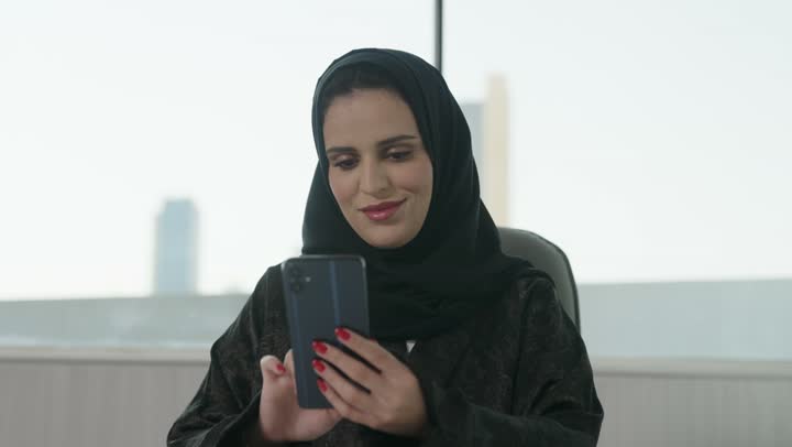 التواصل مع الأهل والأصدقاء عبر الجوال، الدردشة عبر الانترنت, سيدة عربية خليجية سعودية ترتدي العباءة والحجاب تحمل بيدها هاتف محمول , استخدام الأجهزة الحديثة والمتطورة، تصفح وسائل التواصل الاجتماعي.