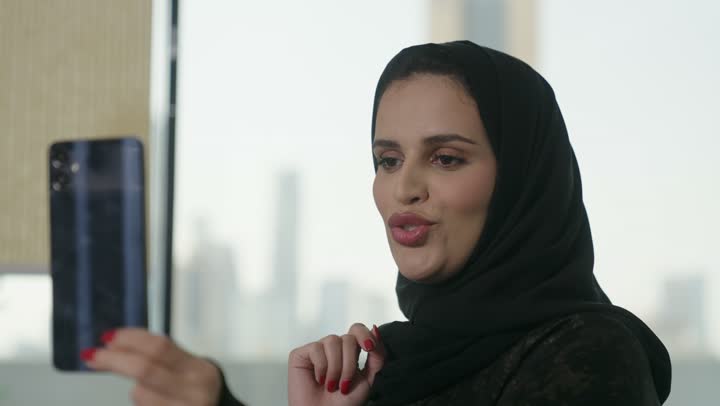 Making a video call via mobile, chatting online, an Arab Gulf Saudi woman wearing an abaya and hijab holding a mobile phone, using modern and advanced devices, browsing social media.