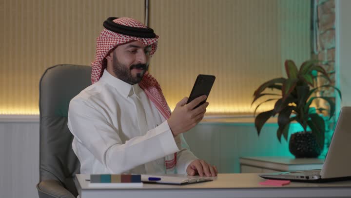 العمل على جهاز الحاسوب المحمول ،الدردشة عبر الانترنت, التواصل مع الأهل والأصدقاء عبر الجوال، رجل عربي خليجي سعودي يرتدي الزي التقليدي والشماغ يحمل بيده هاتف محمول ,  استخدام الأجهزة الحديثة والمتطورة، تصفح وسائل التواصل الاجتماعي