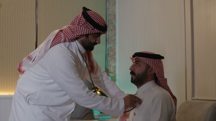 العمل في القطاع الصحي في السعودية، القيام بالرعاية الصحية استخدام سماعة الطبيب لسماع نبضات القلب،  والاهتمام بصحة المرضى، طبيب عربي خليجي سعودي يرتدي عباءة ومئزر أبيض يقوم بفحص رجل سعودي عبر سماعة الأذن،الاهتمام بالصحة الجسدية