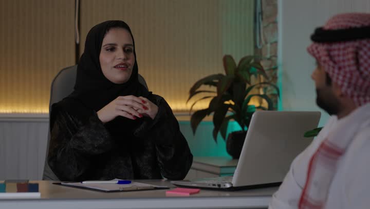 Dialogue and discussion among colleagues, the comfortable work environment in Saudi companies, exchanging experiences and information to achieve goals, two Arab Gulf Saudi employees working in their office within the company headquarters, using computers and technology in the field of business.