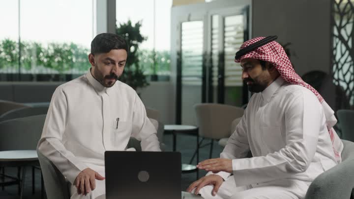 استخدام الحاسوب المحمول والتقنية في مجال الاعمال، الحوار و النقاش بين الزملاء، بيئة العمل المريحة في الشركات السعودية، تبادل الخبرات والمعلومات لتحقيق الاهداف, موظفان عربيان خليجيان سعوديان يعملان في مكتبهم داخل مقر الشركة