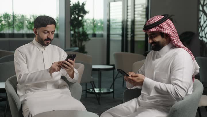 استخدام الجوال والتقنية في مجال الاعمال، الحوار و النقاش بين الزملاء، بيئة العمل المريحة في الشركات السعودية، تبادل الخبرات والمعلومات لتحقيق الاهداف, موظفان عربيان خليجيان سعوديان يعملان في مكتبهم داخل مقر الشركة