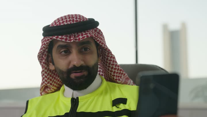 استخدام الأجهزة التقنية الحديثة، العمل في القطاع الهندسي, مهندس عربي خليجي سعودي يرتدي الزي التقليدي وسترة الحماية يحمل الهاتف المحمول يقوم بإجراء مكالمة فيديو، المهن والوظائف الشبابية في السعودية