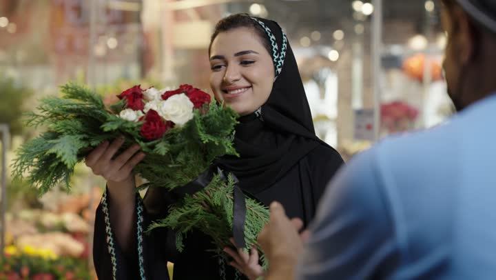 متاجر ومحلات بيع الورود والزهور الطبيعية للأعياد والمناسبات, امرأة عربية خليجية سعودية ترتدي العباءة والحجاب تقوم بشراء باقة ورد احمر طبيعي من البائع , ايماءات تدل على الفرح والسعاده