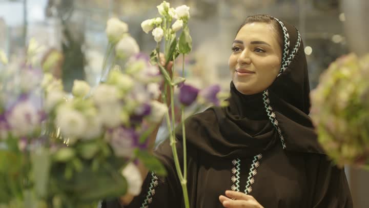 إنتقاء الباقة الأجمل من الورود، إيماءات الإعجاب بالروائح العطرية الزكية للورد، محلات ومتاجر بيع وشراء باقات الورود والهدايا في الأعياد والمناسبات السعيدة، شابة عربية خليجية سعودية ترتدي عباءة مطرزة عصرية تقوم بشم رائحة الزهور