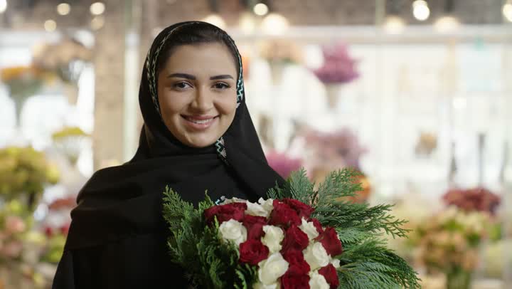 شراء الورود والزهور الطبيعية للأعياد والمناسبات من المحلات والمتاجر, امرأة عربية خليجية سعودية ترتدي العباءة والحجاب تحمل باقة ورد احمر طبيعي بايماءات الفرح والسعاده