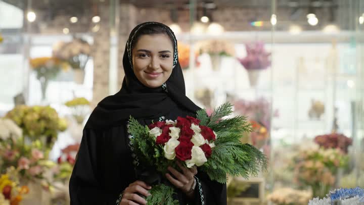 شراء الورود والزهور الطبيعية للأعياد والمناسبات من المحلات والمتاجر, امرأة عربية خليجية سعودية ترتدي العباءة والحجاب تحمل باقة ورد احمر طبيعي بايماءات الفرح والسعاده