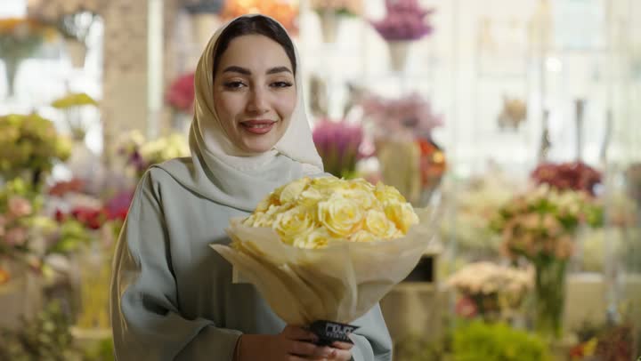 امرأة عربية خليجية سعودية ترتدي العباءة والحجاب تحمل باقة ورد ابيض طبيعي بايماءات الفرح والسعاده، شراء الورود والزهور الطبيعية للأعياد والمناسبات من المحلات والمتاجر,