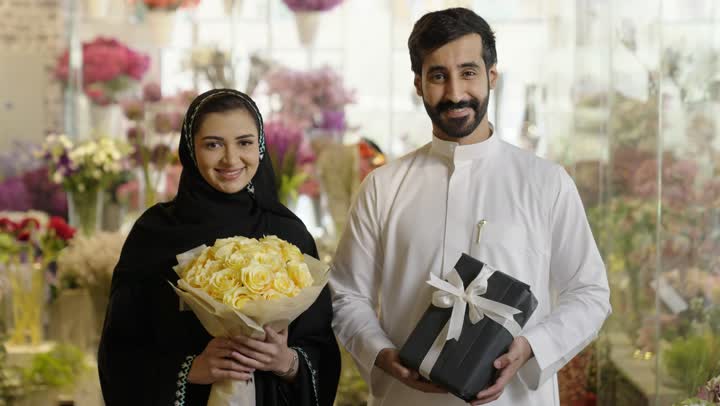 زوجان عربيان خليجيان سعوديان سعيدان يحتفلان بعيد الحب , الزوج يقدم لزوجته باقة ورد , الزوجة تعبر بإيماءات وجه ويدين تدل على الدهشة والفرح , عيد الأم , يوم الفلانتاين , أعياد ومناسبات , عيد الميلاد  .