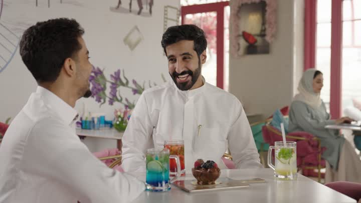 Spending enjoyable time outdoors, entertainment and fun during the holiday, two Arab Gulf men from Saudi Arabia wearing traditional attire sitting in a restaurant with gestures of happiness and joy, sharing hot drinks, coffee, and sweets, exchanging conversations among friends.
