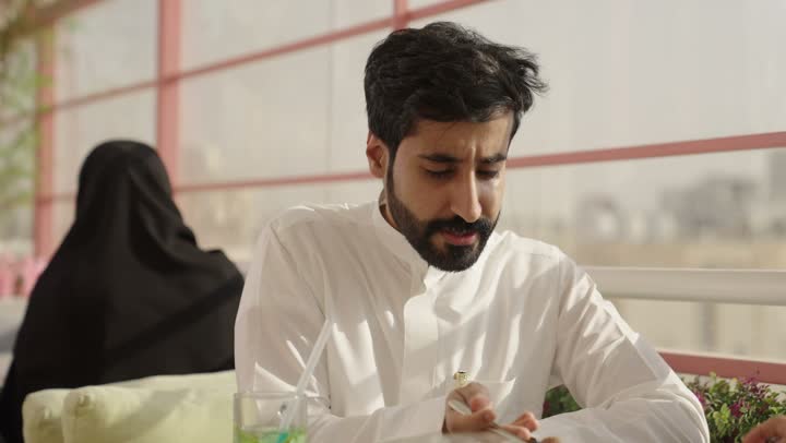 التسلية والترفيه في العطلة، قضاء وقت ممتع في الخارج،رجلان عربيان خليجان سعوديان يرتديان الزي التقليدي يجلسان في المطعم بايماءات السعادة والسرور، مشاركة المشروبات الساخنة والقهوة والحلويات، تبادل الاحاديث بين الأصدقاء