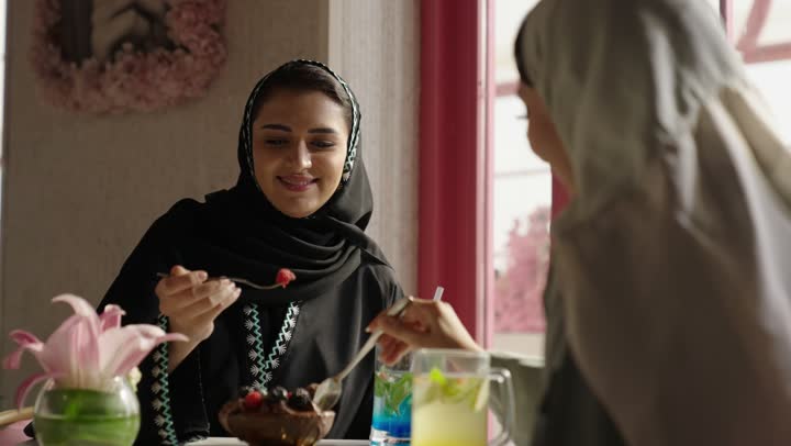 مشاركة المشروبات الساخنة والقهوة والحلويات،التسلية والترفيه في العطلة،قضاء وقت ممتع في الخارج،  صديقتان عربيتان خليجيتان سعوديتان ترتديان العباءه والحجاب تجلسان في المطعم بايماءات السعادة والسرور،  تبادل الاحاديث بين الأصدقاء