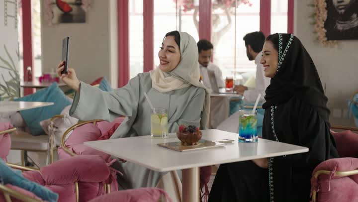 مشاركة المشروبات الساخنة والقهوة والحلويات،التسلية والترفيه في العطلة،قضاء وقت ممتع في الخارج،  صديقتان عربيتان خليجيتان سعوديتان ترتديان العباءه والحجاب تجلسان في المطعم بايماءات السعادة والسرور،  تبادل الاحاديث بين الأصدقاء