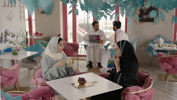 Entertainment and fun during the holiday, sharing hot drinks, coffee, and sweets, spending enjoyable time outdoors, two Arab Gulf Saudi friends wearing abayas and hijabs sitting in a restaurant with gestures of happiness and joy, exchanging conversations among friends.