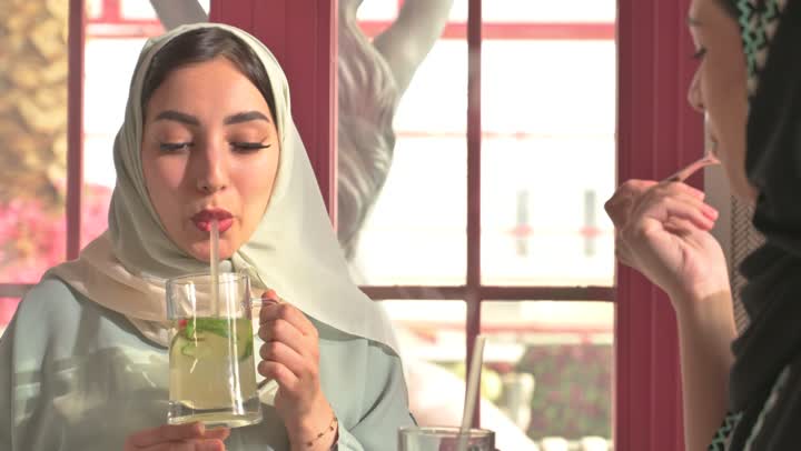 التسلية والترفيه في العطلة، مشاركة المشروبات الساخنة والقهوة والحلويات، قضاء وقت ممتع في الخارج،  صديقتان عربيتان خليجيتان سعوديتان ترتديان العباءه والحجاب تجلسان في المطعم بايماءات السعادة والسرور،  تبادل الاحاديث بين الأصدقاء