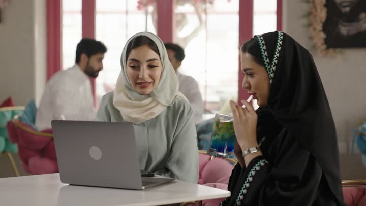 استخدام الحاسوب المحمول ، قضاء وقت ممتع في الخارج، مشاركة المشروبات الساخنة والقهوة والحلويات،التسلية والترفيه في العطلة، صديقتان عربيتان خليجيتان سعوديتان ترتديان العباءه والحجاب تجلسان في المطعم بايماءات السعادة والسرور،  تبادل الاحاديث بين الأصدقاء