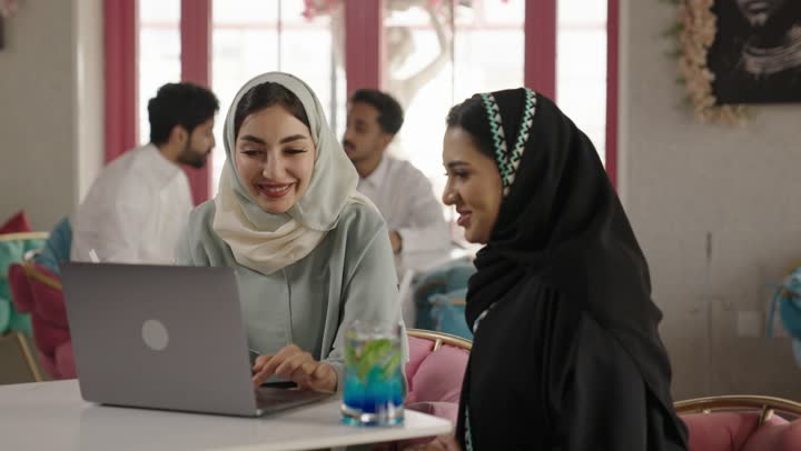 استخدام الحاسوب المحمول ، قضاء وقت ممتع في الخارج، مشاركة المشروبات الساخنة والقهوة والحلويات،التسلية والترفيه في العطلة، صديقتان عربيتان خليجيتان سعوديتان ترتديان العباءه والحجاب تجلسان في المطعم بايماءات السعادة والسرور،  تبادل الاحاديث بين الأصدقاء