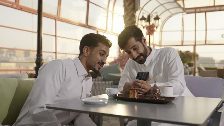 صب القهوه من الدله النحاسية ، قضاء وقت ممتع في الخارج، التسلية والترفيه في العطلة، رجلان عربيان خليجان سعوديان يرتديان الزي التقليدي يجلسان في المطعم بايماءات السعادة والسرور، مشاركة المشروبات الساخنة والقهوة والحلويات، تبادل الاحاديث بين الأصدقاء