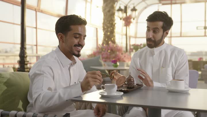 صب القهوه من الدله النحاسية ، قضاء وقت ممتع في الخارج، التسلية والترفيه في العطلة، رجلان عربيان خليجان سعوديان يرتديان الزي التقليدي يجلسان في المطعم بايماءات السعادة والسرور، مشاركة المشروبات الساخنة والقهوة والحلويات، تبادل الاحاديث بين الأصدقاء