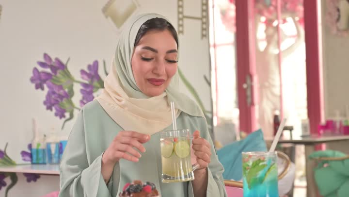 Exchanging conversations between friends, spending enjoyable time outdoors, sharing hot drinks, coffee, and sweets, entertainment and fun during the holiday, two Arab Gulf friends from Saudi Arabia wearing abayas and hijabs sitting in a restaurant with gestures of happiness and joy.