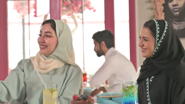 التسلية والترفيه في العطلة، توثيق اللحظات التذكارية باستخدام الجوال ، قضاء وقت ممتع في الخارج، مشاركة المشروبات الساخنة والقهوة والحلويات، صديقتان عربيتان خليجيتان سعوديتان ترتديان العباءه والحجاب تجلسان في المطعم بايماءات السعادة والسرور،  تبادل الاحاديث بين الأصدقاء