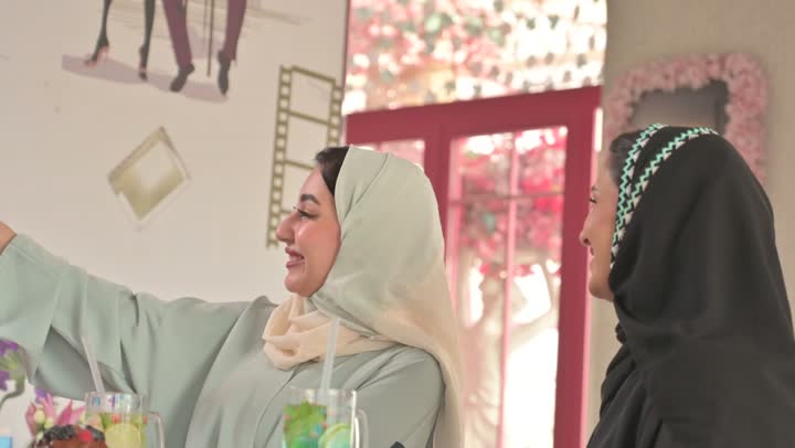 Entertainment and fun during the holiday, documenting memorable moments using a mobile phone, spending enjoyable time outdoors, sharing hot drinks, coffee, and sweets, two Arab Gulf Saudi friends wearing abayas and hijabs sitting in a restaurant with gestures of happiness and joy, exchanging conversations among friends.