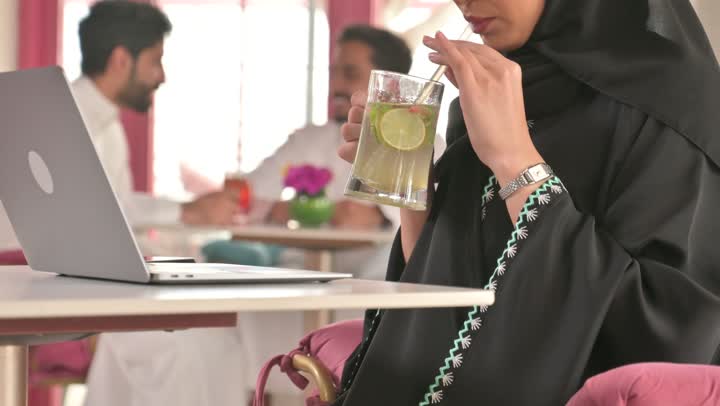 استخدام الحاسوب المحمول ، قضاء وقت ممتع في الخارج، مشاركة المشروبات الساخنة والقهوة والحلويات،التسلية والترفيه في العطلة، صديقتان عربيتان خليجيتان سعوديتان ترتديان العباءه والحجاب تجلسان في المطعم بايماءات السعادة والسرور،  تبادل الاحاديث بين الأصدقاء