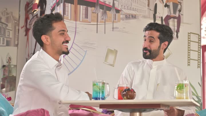 التسلية والترفيه في العطلة، قضاء وقت ممتع في الخارج، رجلان عربيان خليجان سعوديان يرتديان الزي التقليدي يجلسان في المطعم بايماءات السعادة والسرور، مشاركة المشروبات الساخنة والقهوة والحلويات، تبادل الاحاديث بين الأصدقاء