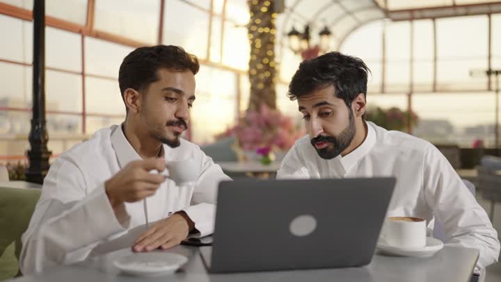 استخدام الحاسوب المحمول ، قضاء وقت ممتع في الخارج، مشاركة المشروبات الساخنة والقهوة والحلويات،التسلية والترفيه في العطلة، رجلان عربيان خليجيان سعوديان  يرتديان الزي التقليدي يجلسان في المطعم بايماءات السعادة والسرور،  تبادل الاحاديث بين الأصدقاء