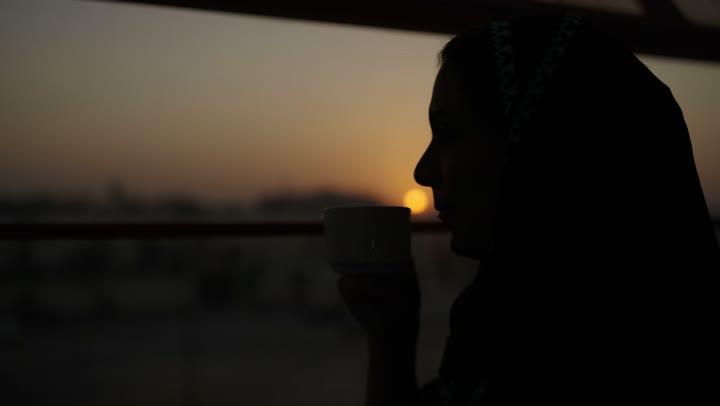 جلسة هادئة مطلة على الميناء البحري وقت غروب الشمس ، تعديل المزاج بشرب الكافيين, الاستمتاع باحتساء المشروب المفضل، سيدة عربية خليجية سعودية ترتدي العباءة تمسك بيدها كوب من القهوة