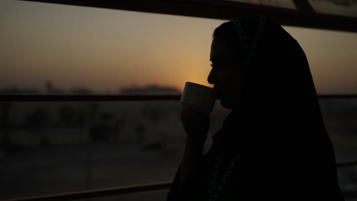جلسة هادئة مطلة على الميناء البحري وقت غروب الشمس ، تعديل المزاج بشرب الكافيين, الاستمتاع باحتساء المشروب المفضل، سيدة عربية خليجية سعودية ترتدي العباءة تمسك بيدها كوب من القهوة