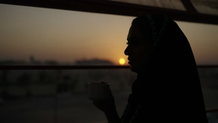 جلسة هادئة مطلة على الميناء البحري وقت غروب الشمس ، تعديل المزاج بشرب الكافيين, الاستمتاع باحتساء المشروب المفضل، سيدة عربية خليجية سعودية ترتدي العباءة تمسك بيدها كوب من القهوة