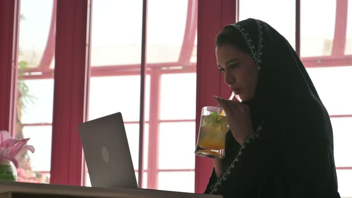 The use of modern and advanced technological devices, managing and organizing business affairs, browsing social media and working remotely in a café, an Arab Gulf Saudi woman wearing an abaya and hijab sitting in a restaurant and using a laptop.
