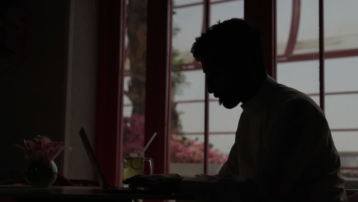 استخدام الأجهزة التقنية الحديثة والمتطورة، ادارة شؤون الاعمال وتنظيمها، تصفح مواقع التواصل الإجتماعي والعمل عن بعد في الكافيه، رجل عربي خليجي سعودي يرتدي الزي التقليدي والشماغ  يجلس في مطعم مظلم  ويستخدم الحاسوب المحمول