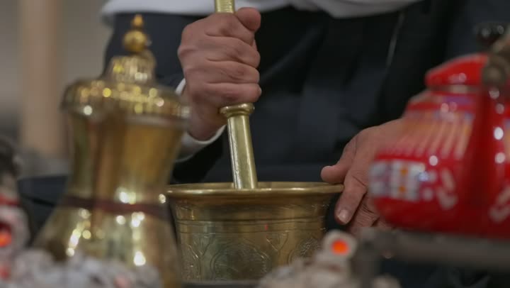 اتباع العادات الأصيلة في صنع المشروبات العربية، استخدام المعاميل لإعداد القهوة بالطريقة التقليدية، محاصيل زراعية وطنية محلية، تصوير مقرب لرجل عربي خليجي سعودي يقوم بطحن حبوب البن في النجر النحاسي