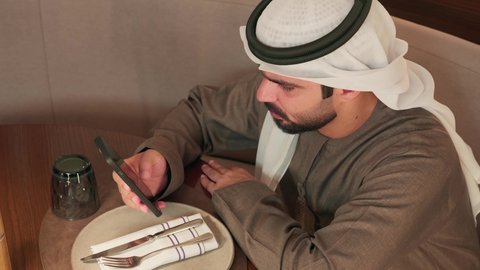 تصفح تطبيقات الهاتف المحمول، الدردشة والتحدث مع الاصدقاء عبر الانترنت، تصوير مقرب لرجل عربي خليجي اماراتي يرتدي الكندورة و الغتره يجلس فى احد المقاهي يقوم بمتابعة اخبار وسائل التواصل الاجتماعي باستخدام الجوال