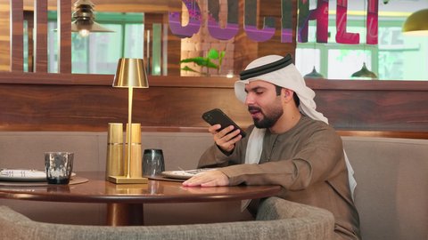 مفهوم ادارة الاعمال عن بعد، التواصل والدردشة مع الأهل والأصدقاء عبر تطبيقات الجوال, رجل عربي خليجي  اماراتي يرتدي  الكندورة و الغتره يجلس فى احدي المقاهي يقوم بإجراء مكالمة  صوتية عبر الهاتف المحمول, استخدام الأجهزة والتقنيات الحديثة المتطورة