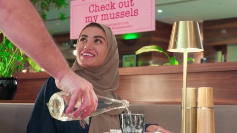 The hospitality and reception, an Arab Gulf Emirati woman wearing an abaya and hijab sits inside the cafe enjoying a pleasant time, restaurants and cafes in the UAE, a close-up shot of a waiter pouring water, attention to detail while serving drinks, a comfortable restaurant atmosphere.