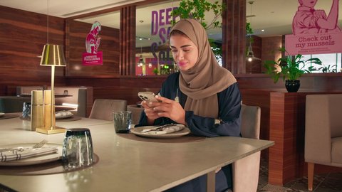 استخدام  الجوال في التسلية و الترفيه ، استعمال البرامج والتطبيقات الالكترونية، امراة عربية خليجية اماراتية محجبة ترتدي العباءة تجلس فى إحدى المقاهي الراقية تستمع بلحظات الهدوء والراحة ، سيده سعودية تقوم بتصفح الهاتف المحمول