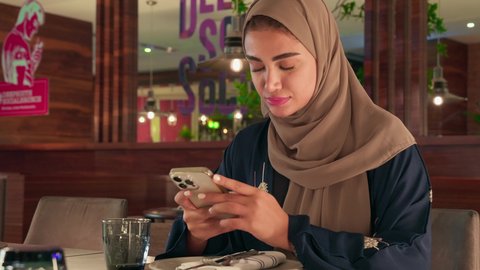 قضاء أوقات ممتعة ومسلية خارج المنزل، استعمال البرامج والتطبيقات الالكترونية، امراة عربية خليجية اماراتية محجبة ترتدي العباءة تجلس فى إحدى المقاهي الراقية تستمع بلحظات الهدوء والراحة ، سيده سعودية تقوم بتصفح الهاتف المحمول