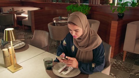 استعمال البرامج والتطبيقات الالكترونية، قضاء أوقات ممتعة ومسلية خارج المنزل، امراة عربية خليجية اماراتية محجبة ترتدي العباءة تجلس فى إحدى المقاهي الراقية تستمع بلحظات الهدوء والراحة ، سيده سعودية تقوم بتصفح الهاتف المحمول