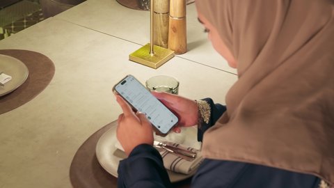 دردشات وحوارات ودية مع الأصحاب، سيده سعودية تقوم بتصفح مواقع التواصل الالكتروني عبىر الهاتف المحمول، استعمال البرامج والتطبيقات الالكترونية، تصوير مقرب من الخلف لامراة عربية خليجية اماراتية محجبة ترتدي العباءة تجلس فى إحدى المقاهي الراقية تستمع بلحظات الهدوء والراحة