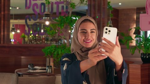 Documenting happy moments, taking commemorative selfies via mobile, a Saudi woman enjoying her time at the cafe, a veiled Emirati Gulf Arab woman wearing an abaya holding a mobile phone and making a victory sign with her hands.