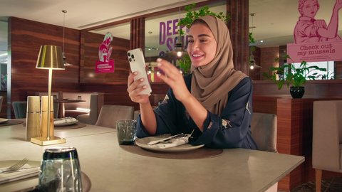 An Arab Gulf woman from the UAE, wearing an abaya and hijab, is sitting in a cafe making a video call on her mobile phone, communicating and chatting with family and friends through mobile applications. A Saudi woman is using modern advanced devices and technologies.