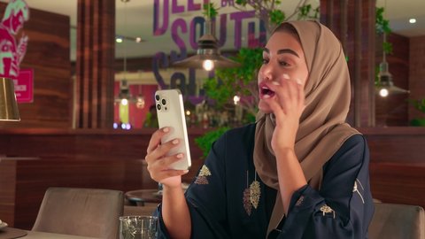 التواصل والدردشة مع الأهل والأصدقاء عبر تطبيقات الجوال,امراه عربيه خليجيه اماراتية ترتدي العباءة و الحجاب تجلس فى احد المقاهي  تقوم بإجراء مكالمة فيديو عبر الهاتف المحمول,  سيده سعودية تستخدم الأجهزة والتقنيات الحديثة المتطورة، تناول المشروب المفضل